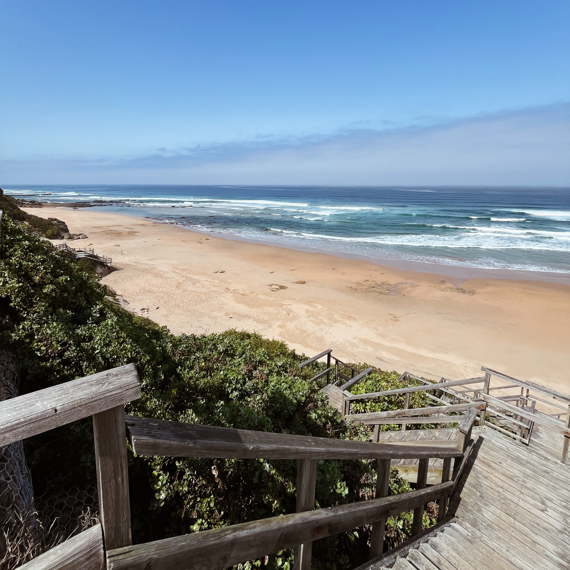 Wooden staircase leading down to a sandy beach with ocean waves and clear blue sky.